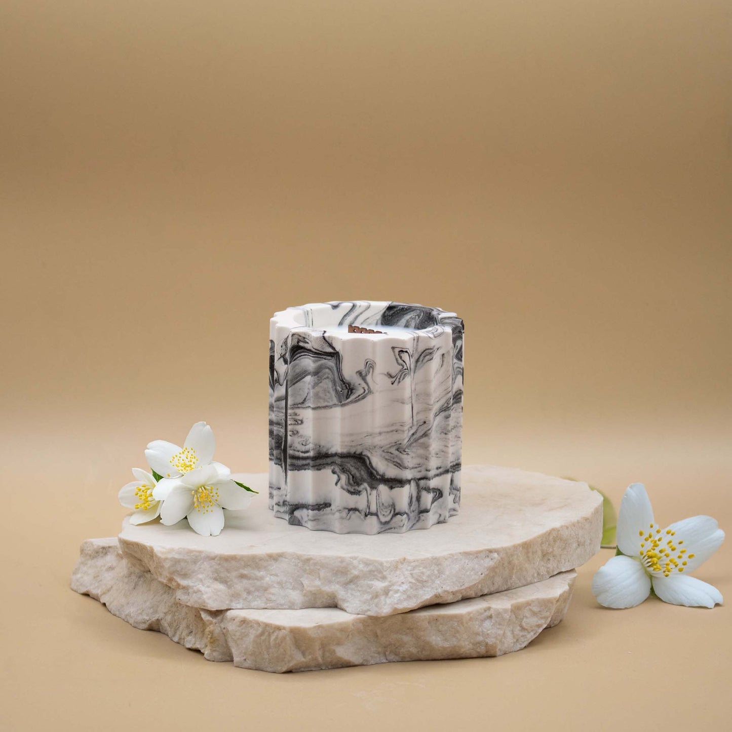Bougie artisanale marbrée dans des tons de blanc et de noir en forme de colonne romaine, posée sur une pierre naturelle, accompagnée de fleurs de jasmin délicates sur un fond beige.