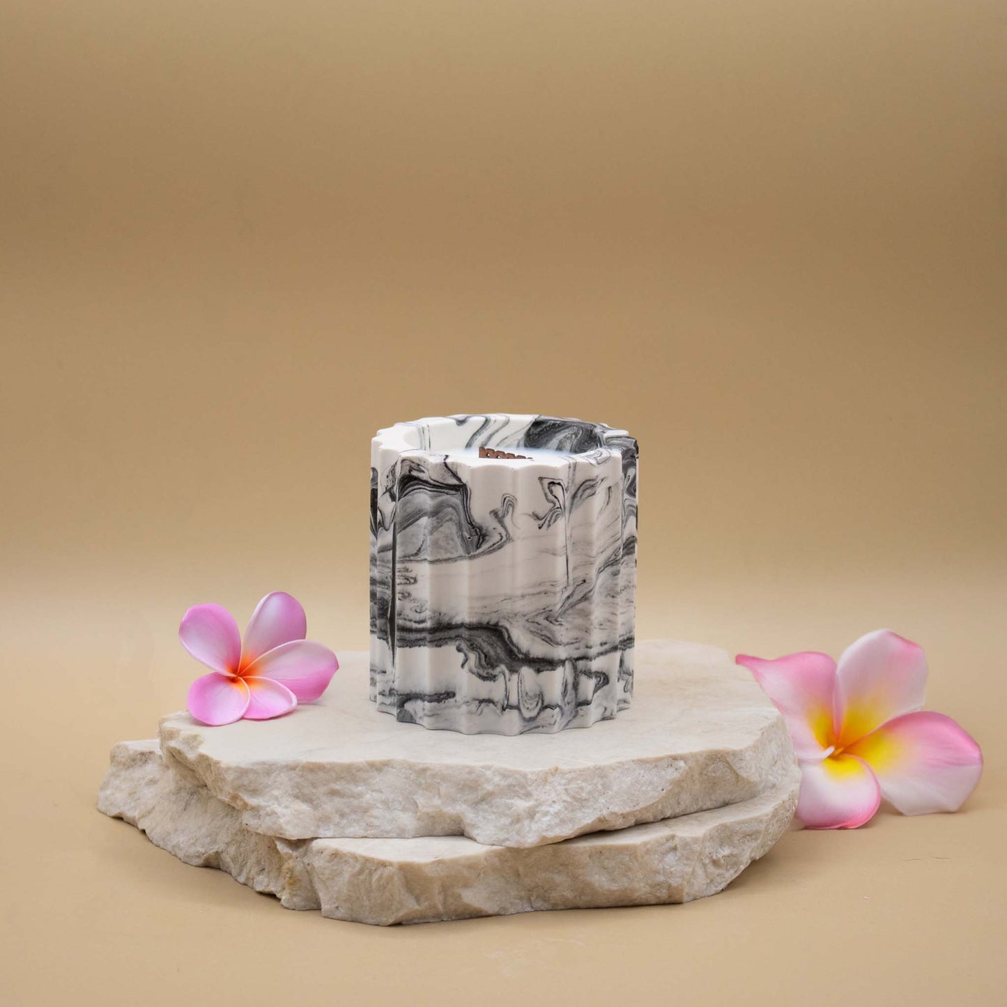 Bougie artisanale marbrée dans des tons de blanc et de noir en forme de colonne romaine, posée sur une pierre naturelle, accompagnée de fleurs exotiques évoquant le monoï, sur un fond beige.