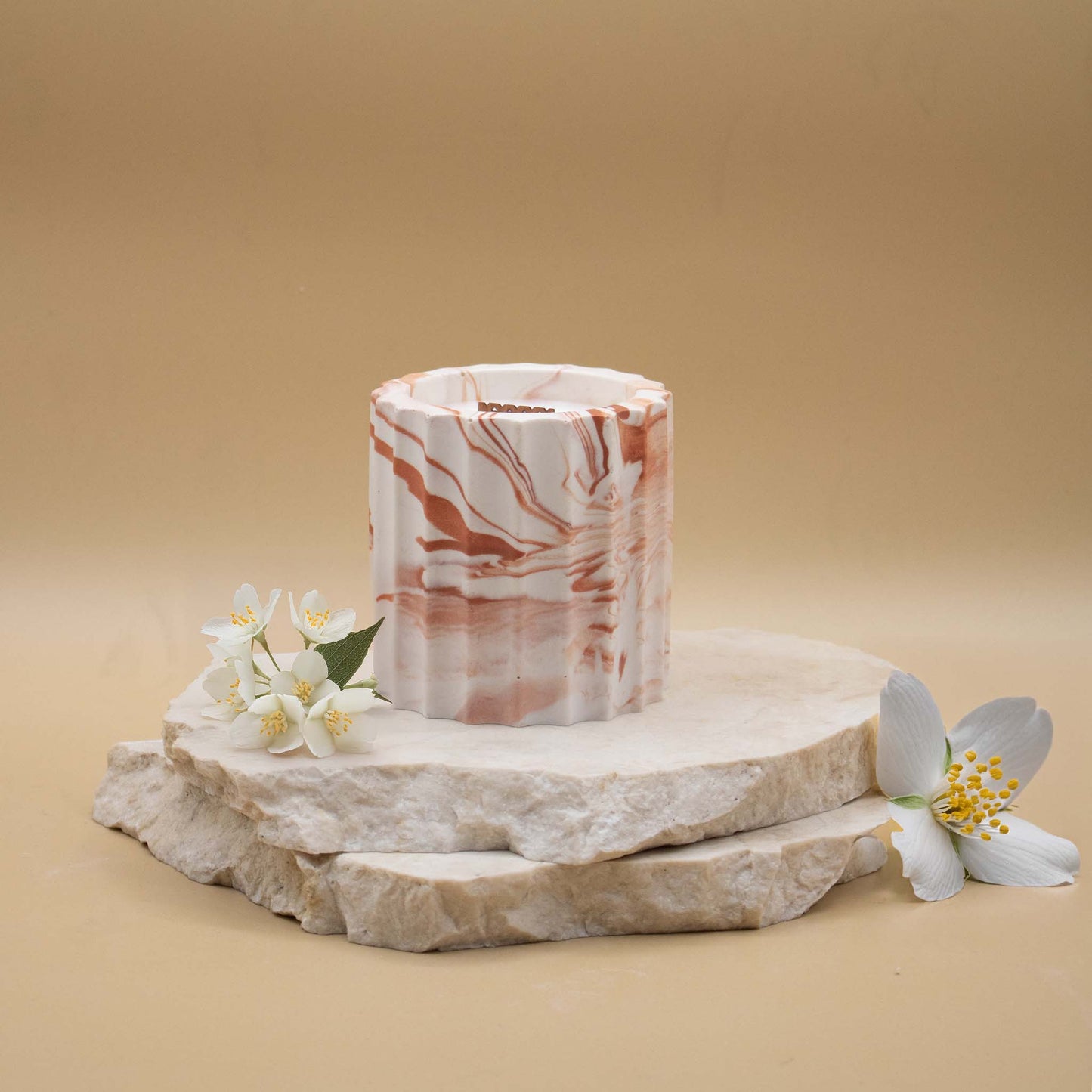 Bougie artisanale marbrée dans des tons de blanc et de terracotta en forme de colonne romaine, posée sur une pierre naturelle, accompagnée de fleurs de jasmin délicates sur un fond beige.