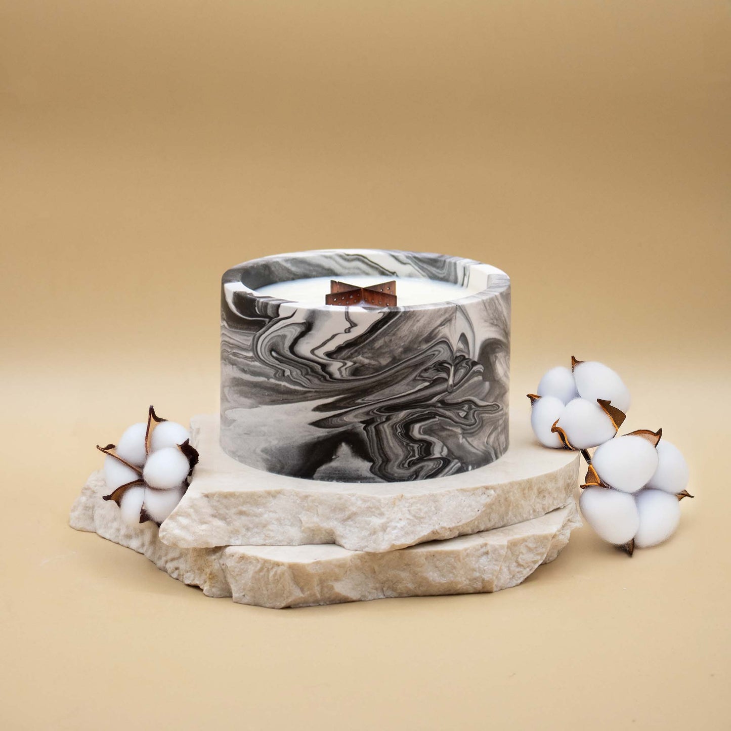Grande bougie ronde artisanale marbrée en noir et blanc avec une mèche en bois, posée sur une pierre naturelle, accompagnée de fleurs délicates représentant la fleur de coton, sur un fond beige