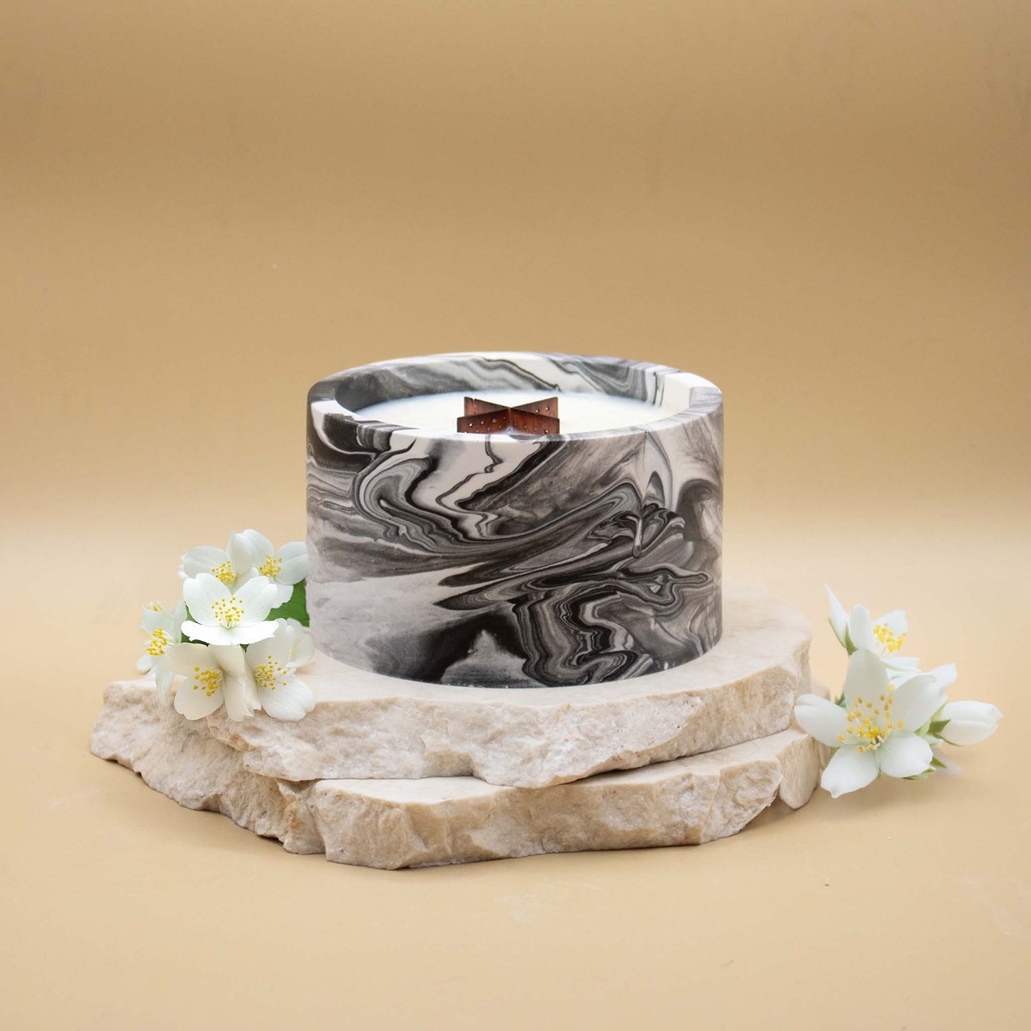 Grande bougie ronde artisanale marbrée en noir et blanc avec une mèche en bois, posée sur une pierre naturelle, accompagnée de fleurs blanches délicates évoquant le jasmin, sur un fond beige.