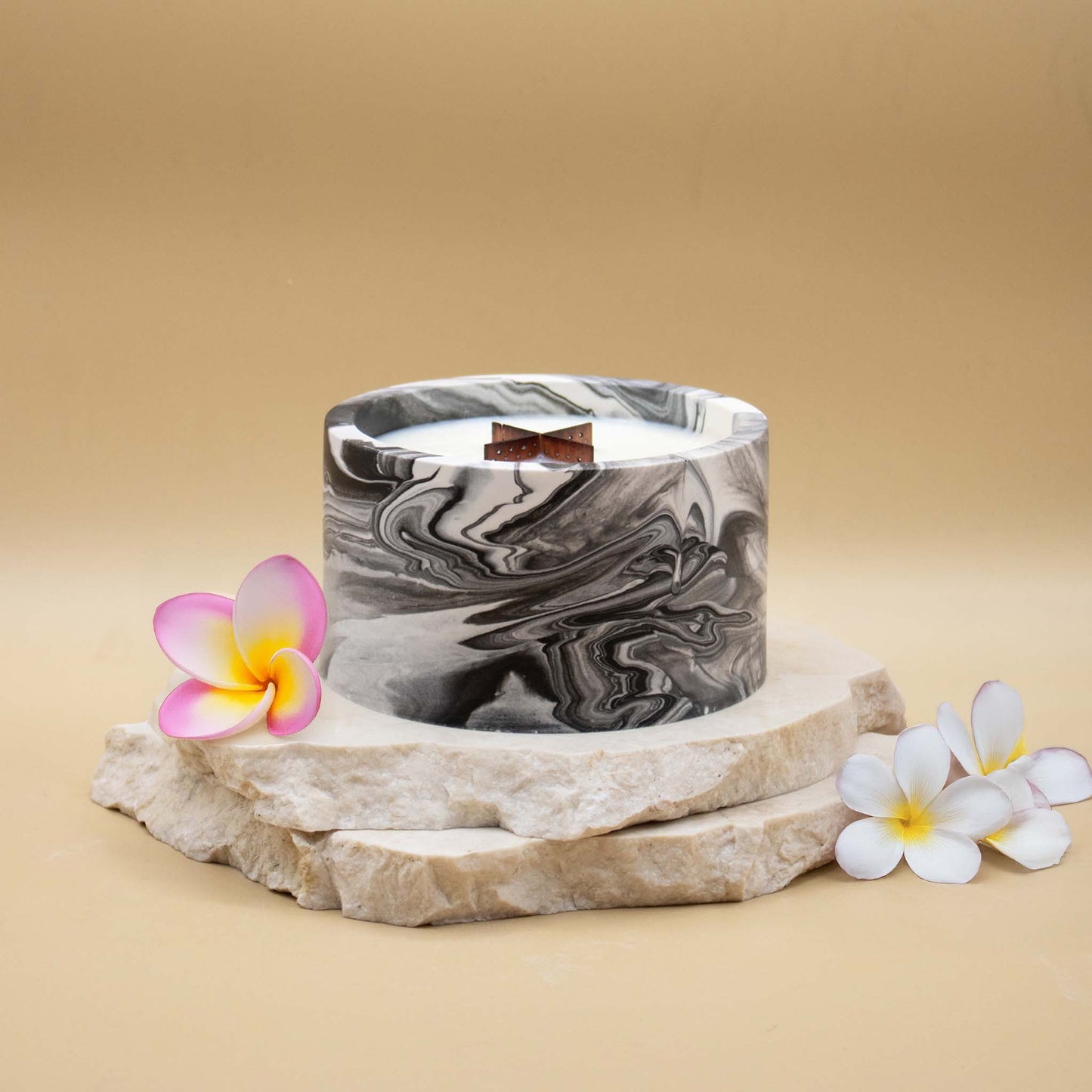 Grande bougie ronde artisanale marbrée en noir et blanc avec une mèche en bois, posée sur une pierre naturelle, accompagnée de fleurs exotiques évoquant le monoï, sur un fond beige.
