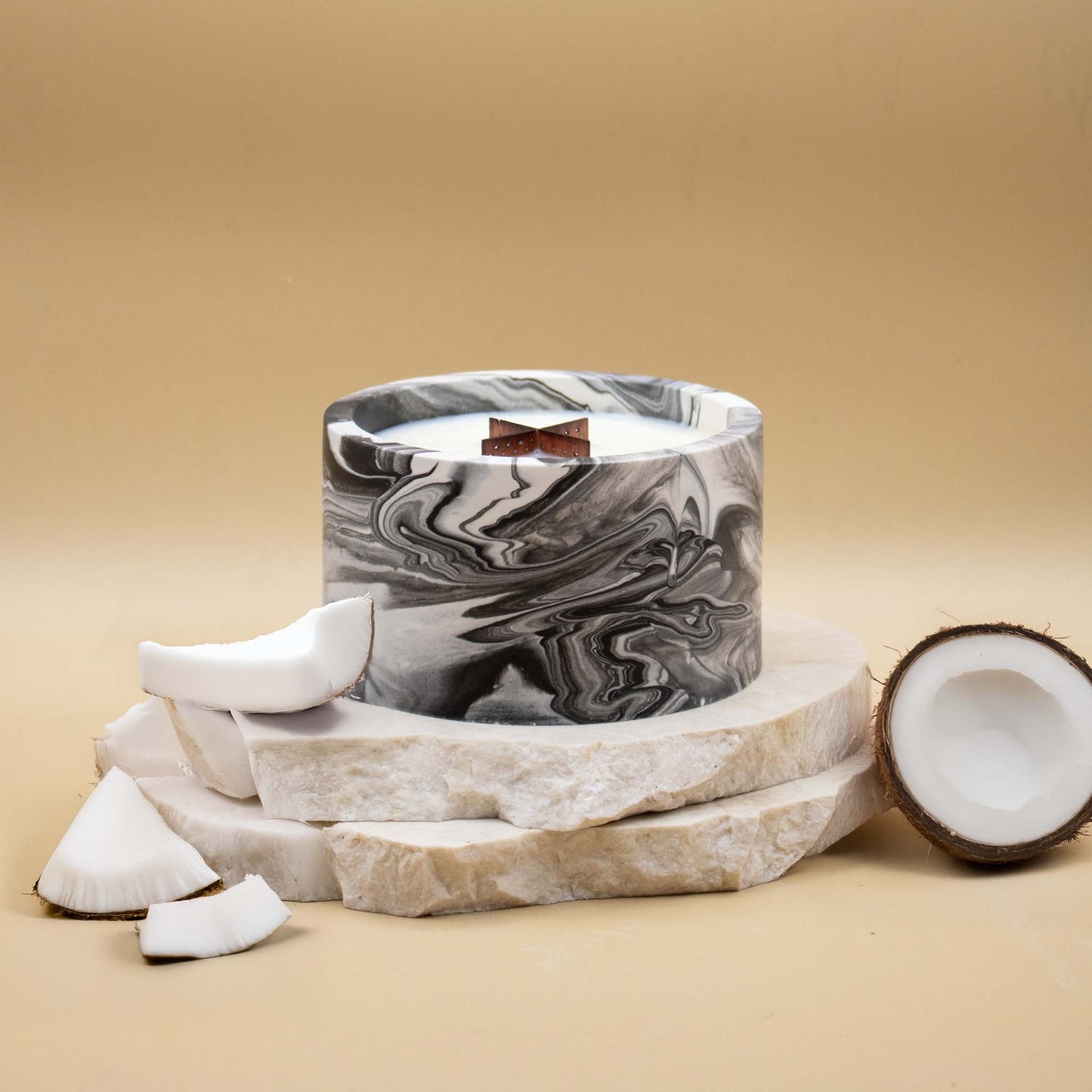 Grande bougie ronde artisanale marbrée en noir et blanc avec une mèche en bois, posée sur une pierre naturelle, accompagnée d'éléments évoquant la noix de coco, sur un fond beige.