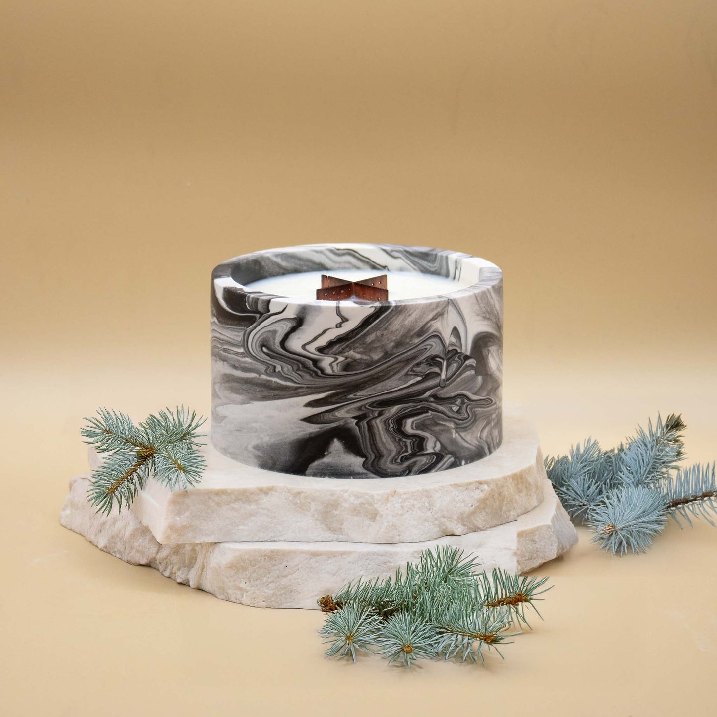 Grande bougie ronde artisanale marbrée en noir et blanc avec une mèche en bois, posée sur une pierre naturelle, accompagnée de branches ou aiguilles de sapin, sur un fond beige.