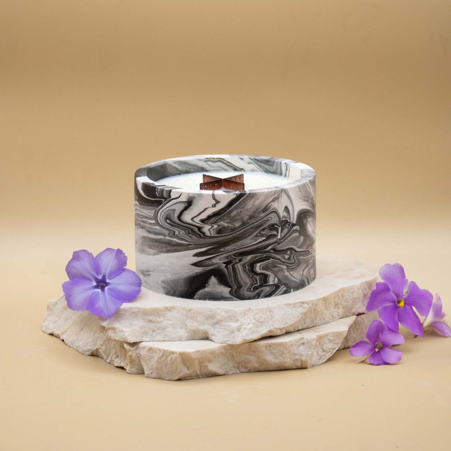 Grande bougie ronde artisanale marbrée en noir et blanc avec une mèche en bois, posée sur une pierre naturelle, accompagnée de fleurs violettes évoquant la violette, sur un fond beige.