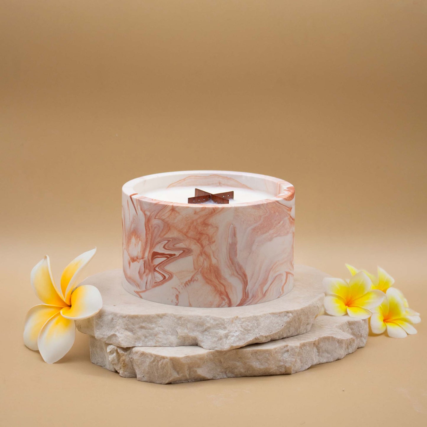 Grande bougie ronde artisanale marbrée en terracotta avec une mèche en bois, posée sur une pierre naturelle, accompagnée de fleurs exotiques évoquant le monoï, sur un fond beige.
