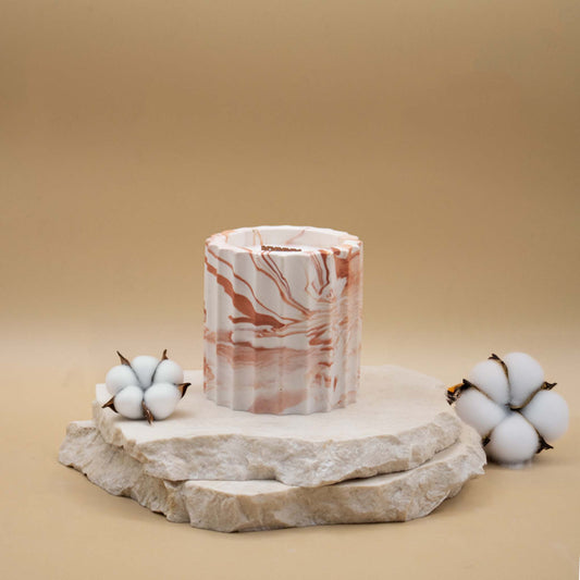Bougie artisanale marbrée dans des tons de blanc et de terracotta en forme de colonne romaine, posée sur une pierre naturelle, accompagnée de fleurs délicates représentant la fleur de coton, sur un fond beige