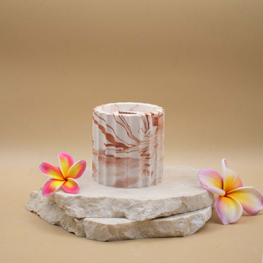 Bougie artisanale marbrée dans des tons de blanc et de terracotta en forme de colonne romaine, posée sur une pierre naturelle, accompagnée de fleurs exotiques évoquant le monoï, sur un fond beige.