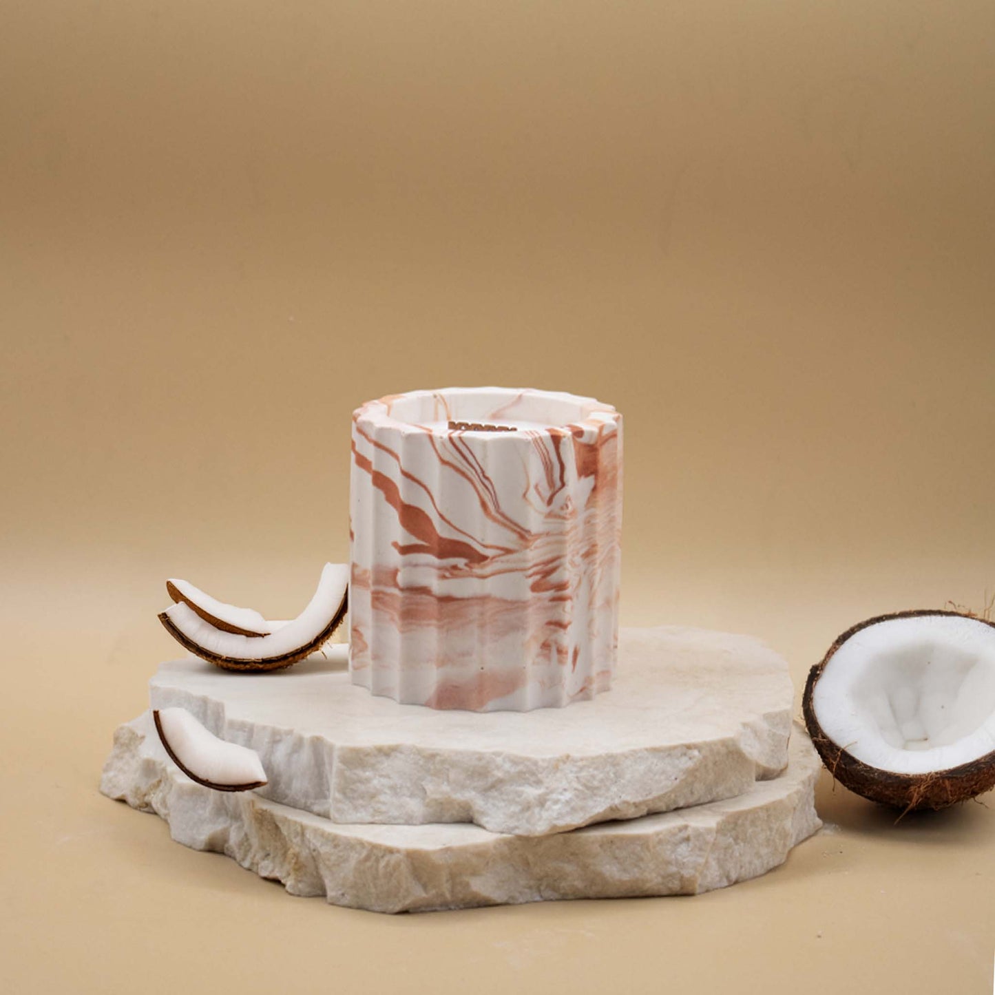 Bougie artisanale marbrée dans des tons de blanc et de terracotta en forme de colonne romaine, posée sur une pierre naturelle, accompagnée d'éléments évoquant la noix de coco, sur un fond beige.