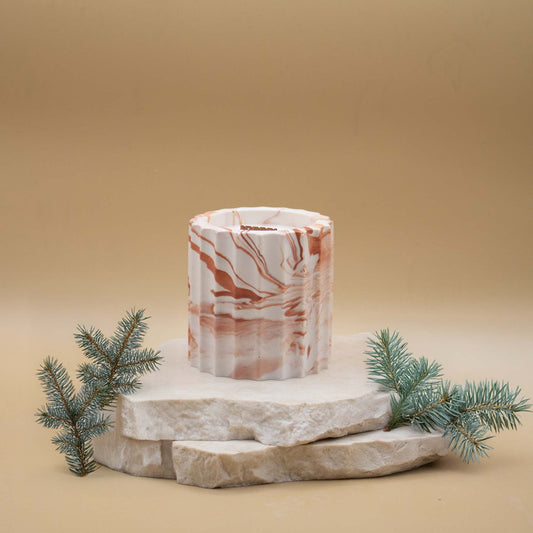 Bougie artisanale marbrée dans des tons de blanc et de terracotta en forme de colonne romaine, posée sur une pierre naturelle, accompagnée de branches de sapin, sur un fond beige.