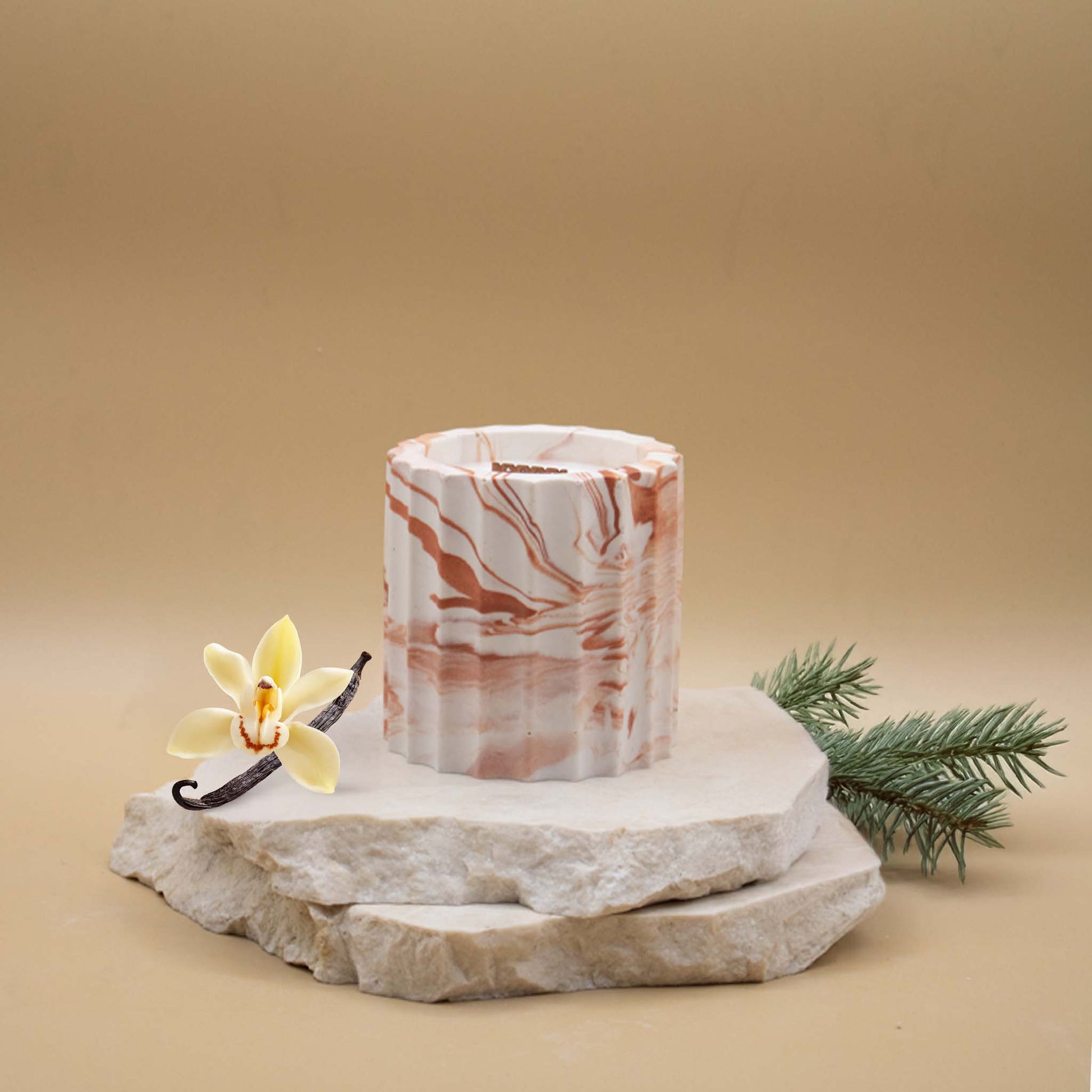 Bougie artisanale marbrée dans des tons de blanc et de terracotta en forme de colonne romaine, posée sur une pierre naturelle, accompagnée d'éléments évoquant la vanille boisée, sur un fond beige.