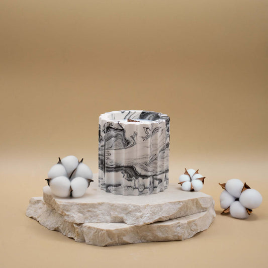 Bougie artisanale marbrée dans des tons de blanc et de noir en forme de colonne romaine, posée sur une pierre naturelle, accompagnée de fleurs délicates représentant la fleur de coton, sur un fond beige