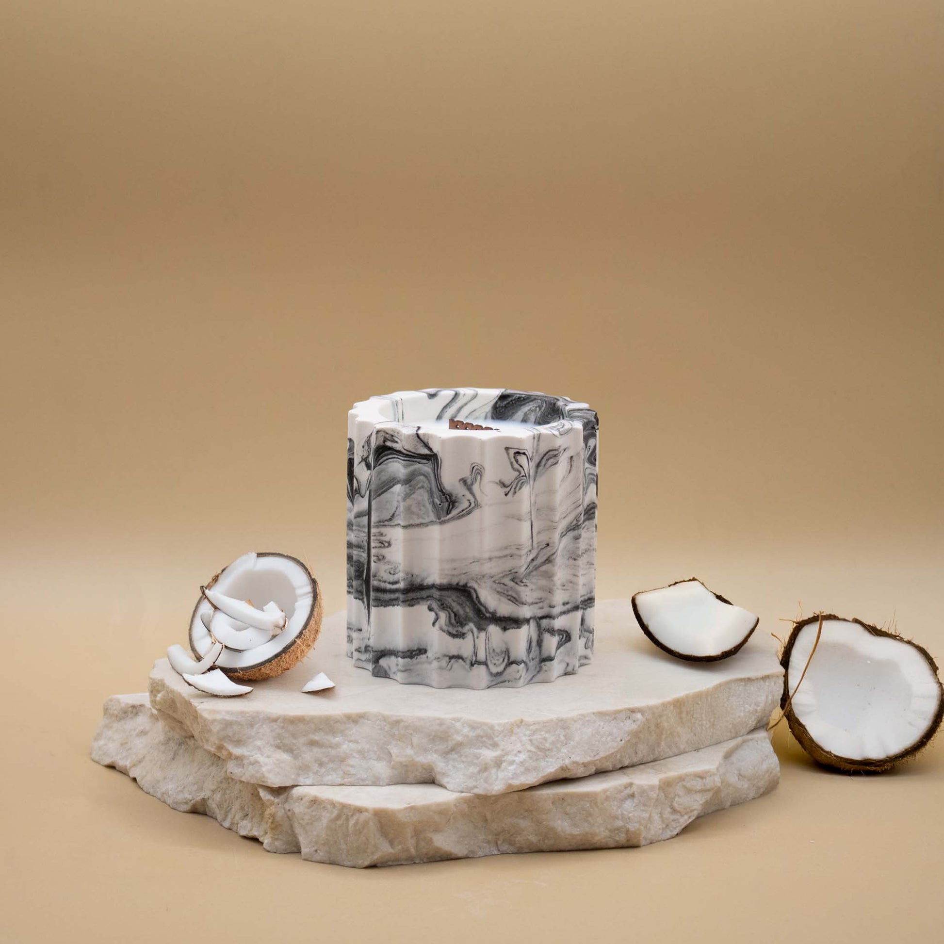 Bougie artisanale marbrée dans des tons de blanc et de noir en forme de colonne romaine, posée sur une pierre naturelle, accompagnée d'éléments évoquant la noix de coco, sur un fond beige.