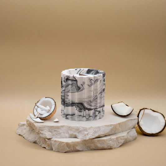 Bougie artisanale marbrée dans des tons de blanc et de noir en forme de colonne romaine, posée sur une pierre naturelle, accompagnée d'éléments évoquant la noix de coco, sur un fond beige.