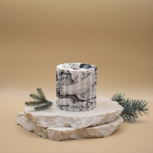 Bougie artisanale marbrée dans des tons de blanc et de noir en forme de colonne romaine, posée sur une pierre naturelle, accompagnée de branches de sapin, sur un fond beige.
