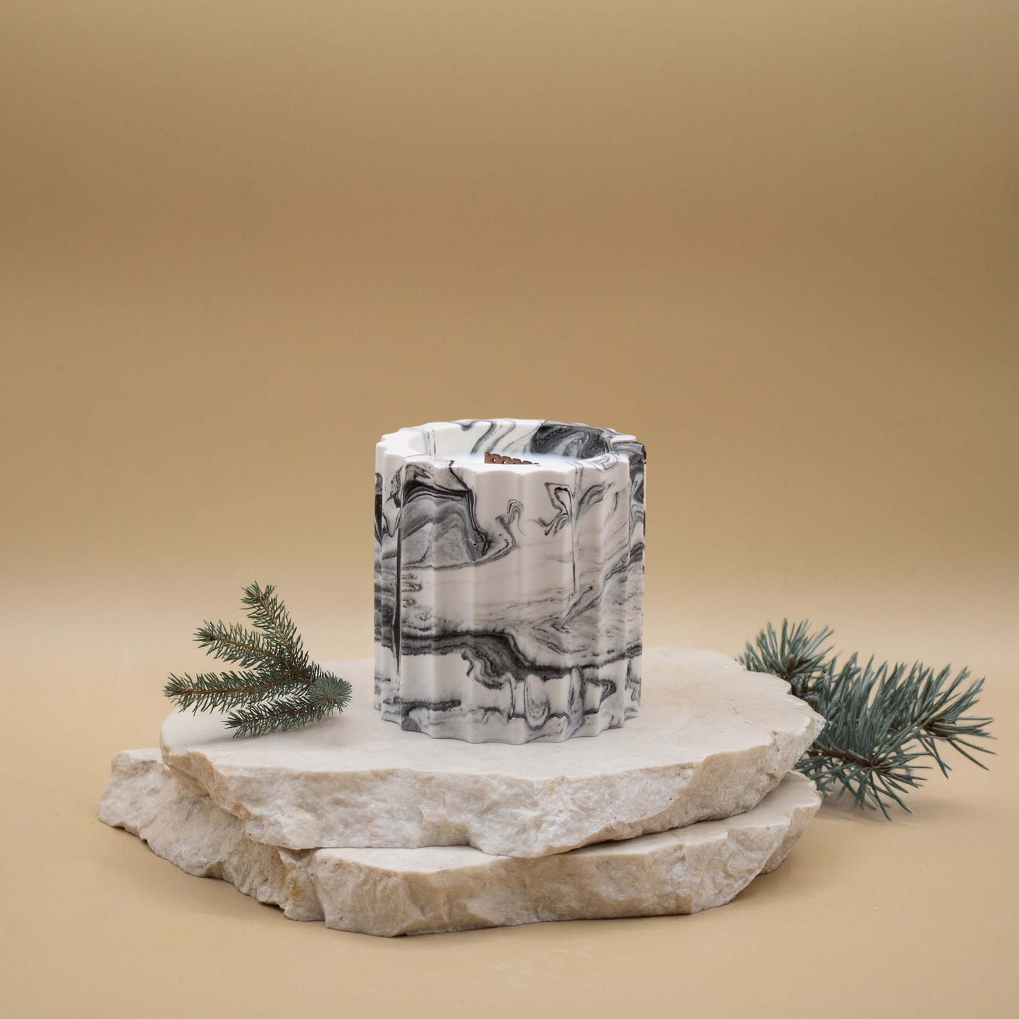 Bougie artisanale marbrée dans des tons de blanc et de noir en forme de colonne romaine, posée sur une pierre naturelle, accompagnée de branches de sapin, sur un fond beige.