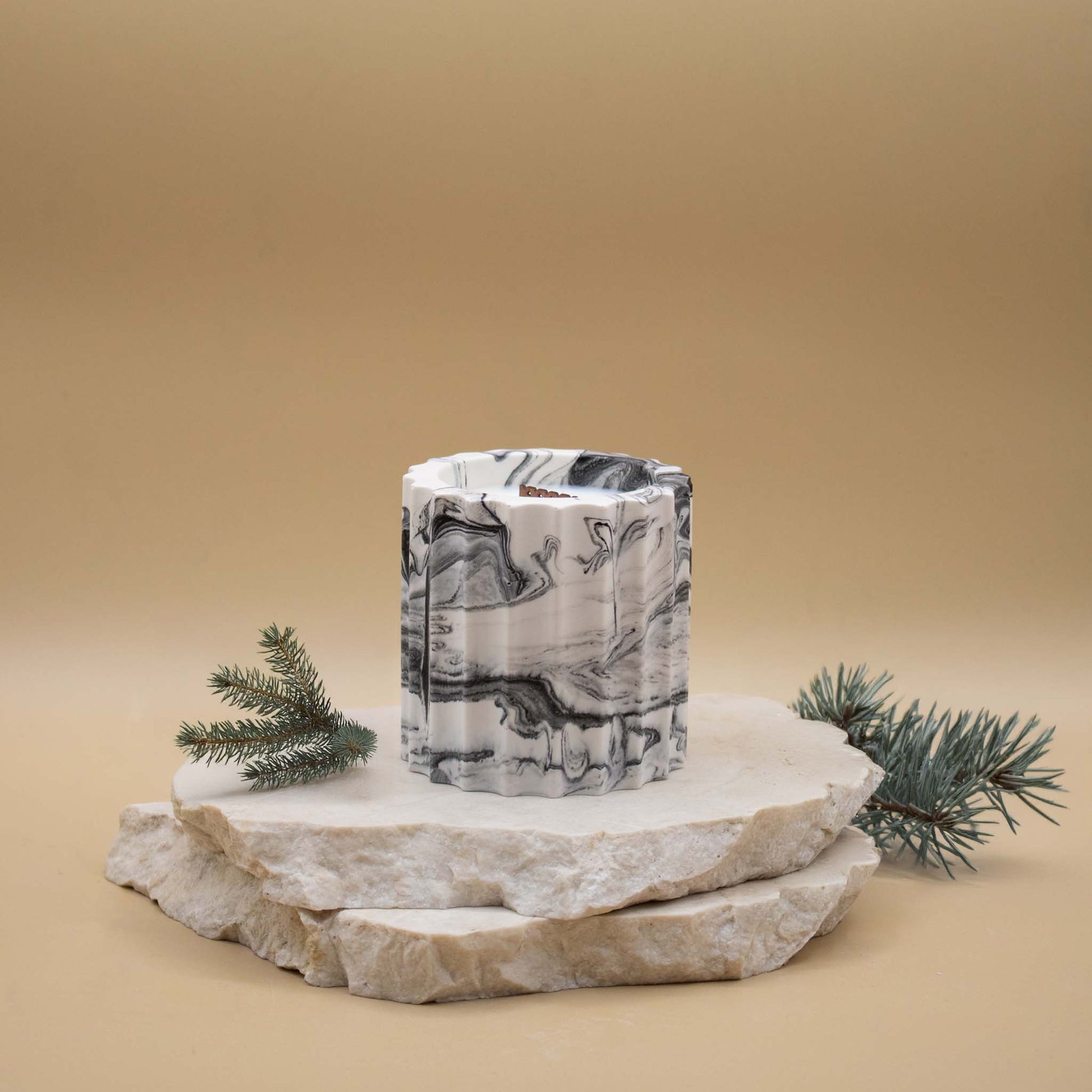 Bougie artisanale marbrée dans des tons de blanc et de noir en forme de colonne romaine, posée sur une pierre naturelle, accompagnée de branches de sapin, sur un fond beige.