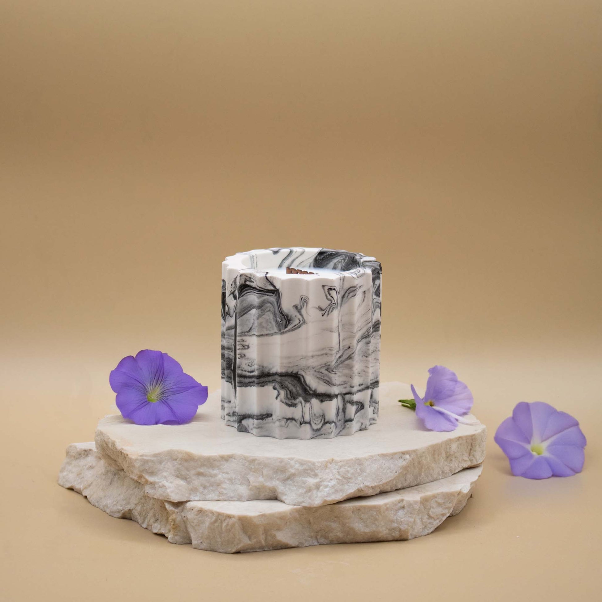 Bougie artisanale marbrée dans des tons de blanc et de noir en forme de colonne romaine, posée sur une pierre naturelle, accompagnée de fleurs évoquant la violette, sur un fond beige.