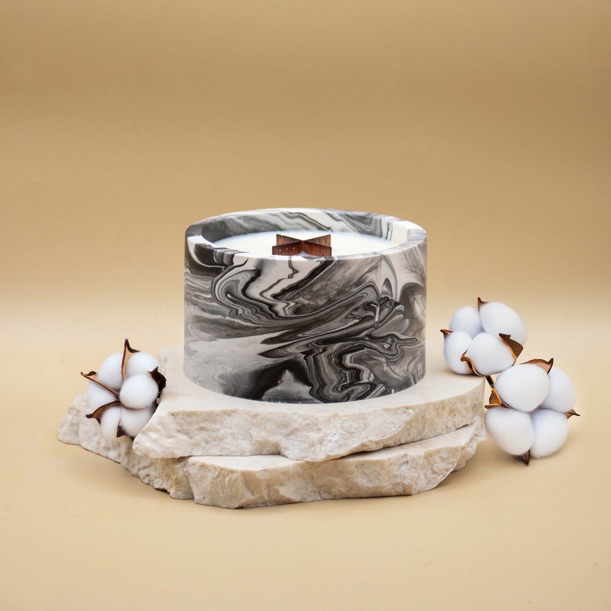 Grande bougie ronde artisanale marbrée en noir et blanc avec une mèche en bois, posée sur une pierre naturelle, accompagnée de fleurs délicates représentant la fleur de coton, sur un fond beige
