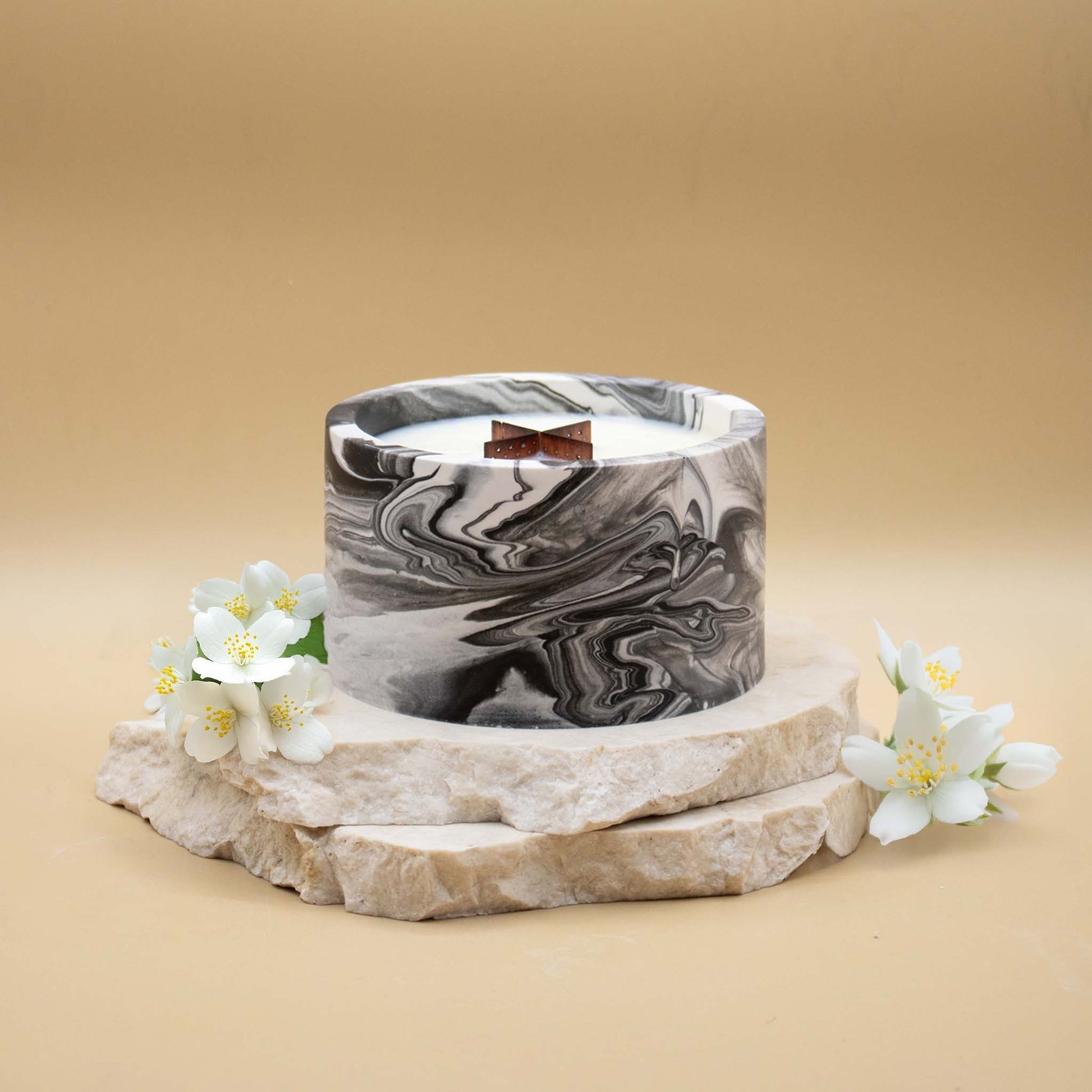 Grande bougie ronde artisanale marbrée en noir et blanc avec une mèche en bois, posée sur une pierre naturelle, accompagnée de fleurs blanches délicates évoquant le jasmin, sur un fond beige.