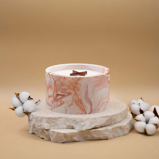 Grande bougie ronde artisanale marbrée en terracotta avec une mèche en bois, posée sur une pierre naturelle, accompagnée de fleurs délicates représentant la fleur de coton, sur un fond beige