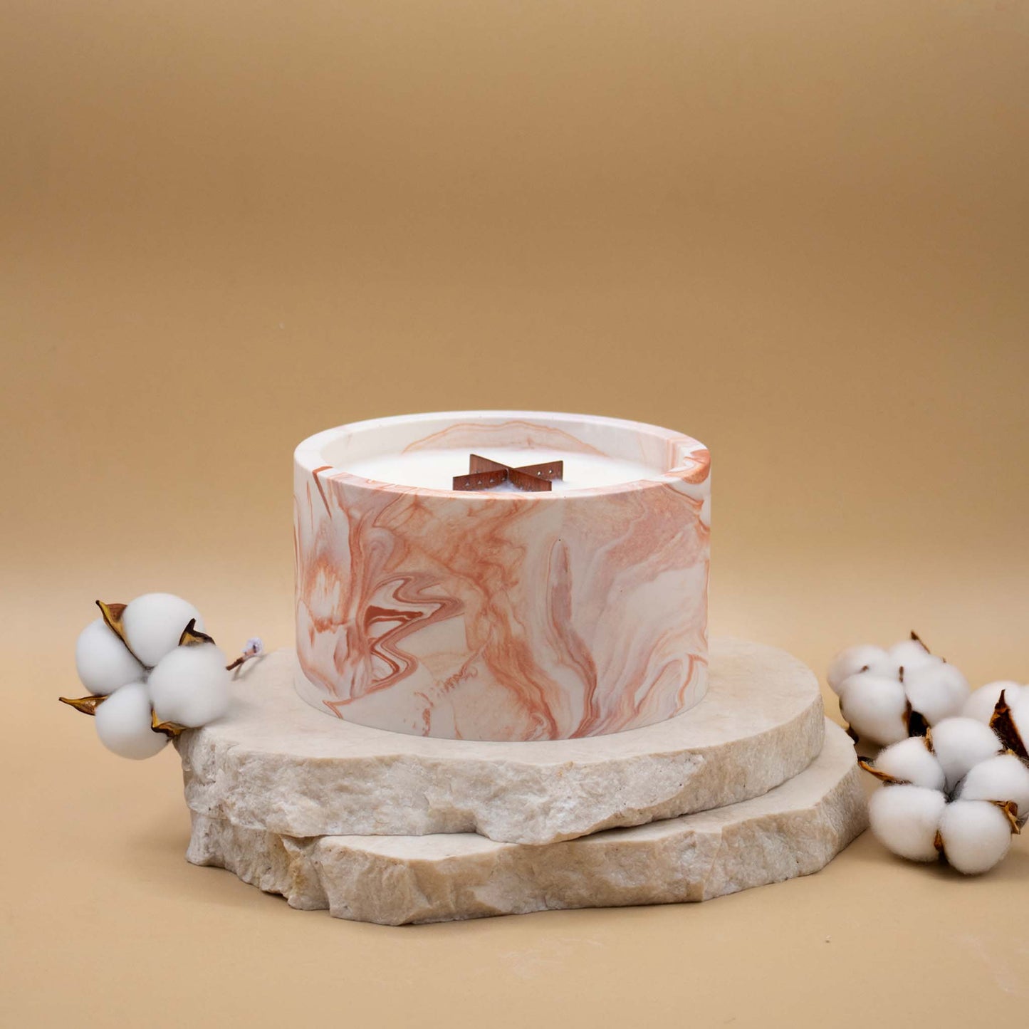 Grande bougie ronde artisanale marbrée en terracotta avec une mèche en bois, posée sur une pierre naturelle, accompagnée de fleurs délicates représentant la fleur de coton, sur un fond beige