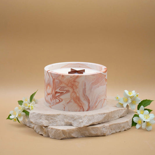 Grande bougie ronde artisanale marbrée en terracotta avec une mèche en bois, posée sur une pierre naturelle, accompagnée de fleurs blanches délicates évoquant le jasmin, sur un fond beige.
