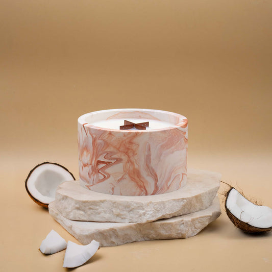 Grande bougie ronde artisanale marbrée en terracotta avec une mèche en bois, posée sur une pierre naturelle, accompagnée d'éléments évoquant la noix de coco, sur un fond beige.