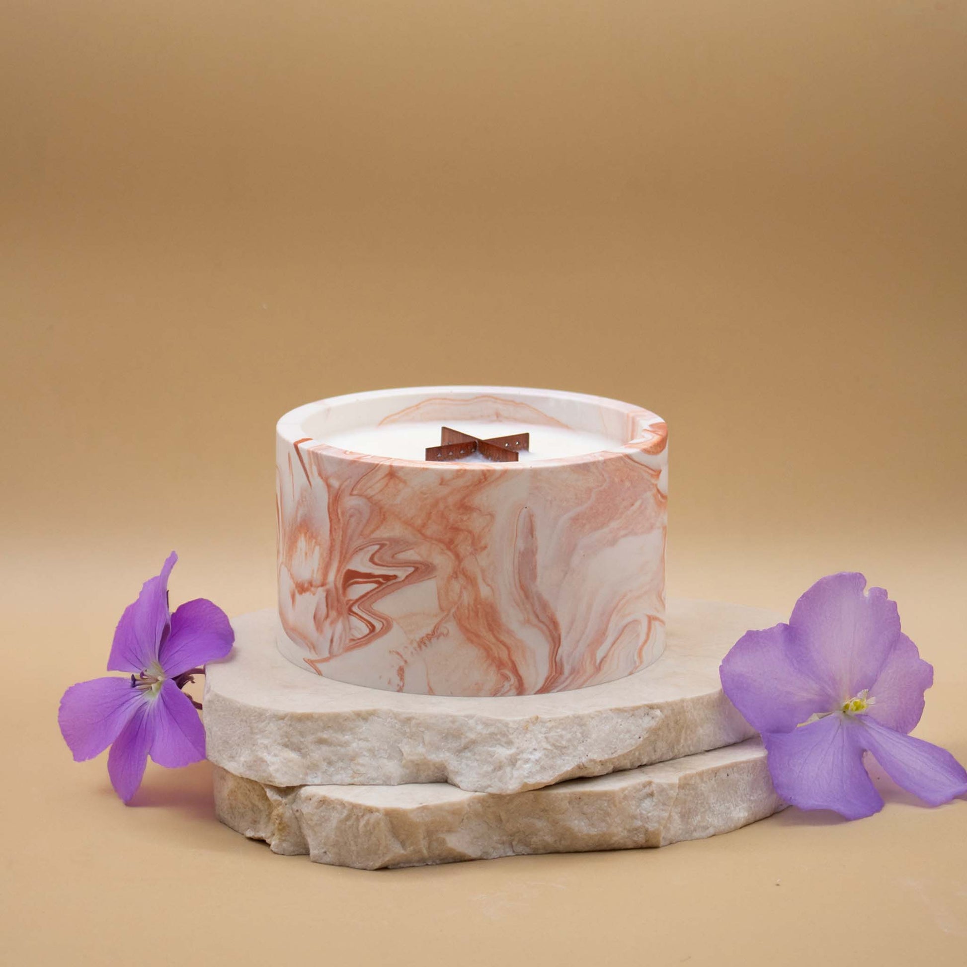 Grande bougie ronde artisanale marbrée en terracotta avec une mèche en bois, posée sur une pierre naturelle, accompagnée de fleurs violettes évoquant la violette, sur un fond beige.